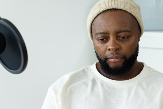 African man sitting in home studio recording with mic pop filter wearing knit beanie cream shirt