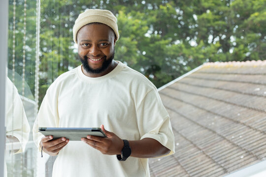 Adult African man standing holding tablet by wet glass strands, wearing knit beanie and black watch