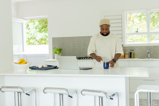 Man wearing beanie stirring dark blue mug with spoon at kitchen island with laptop smartphone