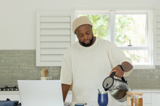 Adult African American man pouring coffee from glass carafe into mug at kitchen counter, laptop