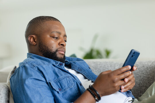 African American adult reclining on sofa at home using smartphone, wearing denim shirt, bracelet
