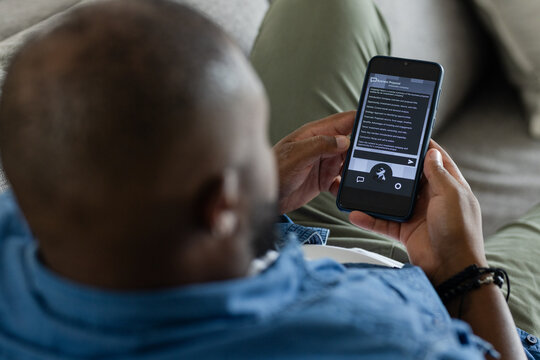 African American man reclining on pale sofa at home, in denim shirt and reading smartphone article
