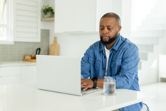 African American man wearing denim shirt working on laptop at kitchen island, glass, copy space