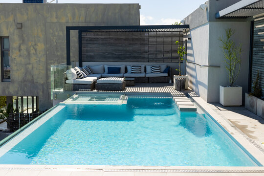 Rectangular turquoise pool is glistening on rooftop terrace with pergola shadows and wicker sofa