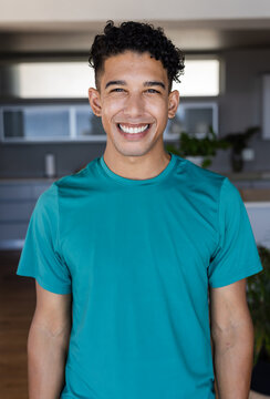 African American man smiling, standing in kitchen in teal shirt hoop earrings with cabinets window