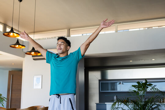 African American man standing with arms raised and eyes closed at home near pendant lights, plants