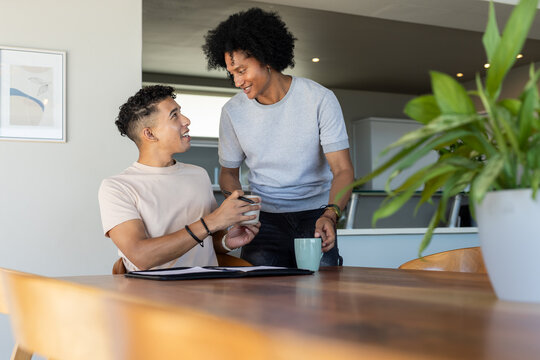 African American male friends sitting at home dining table holding mugs near black binder and plant