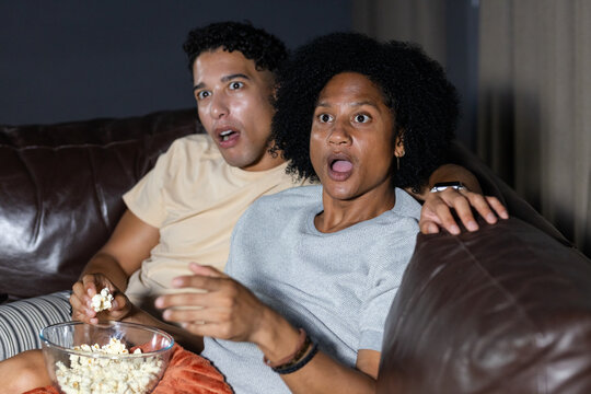 African American male friends leaning forward, watching TV in living room, holding popcorn bowl