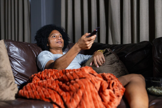 Adult African man reclining on leather couch at home holding TV remote under orange throw blanket