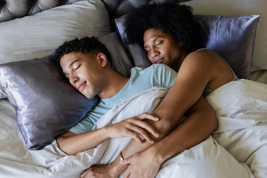 African American couple spooning and holding each other in bed with white duvet, gold ring visible