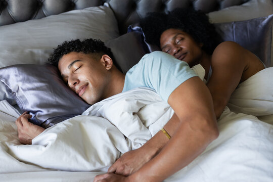 African American men cuddling and sleeping in bed with tufted headboard, satin pillows, smartphone