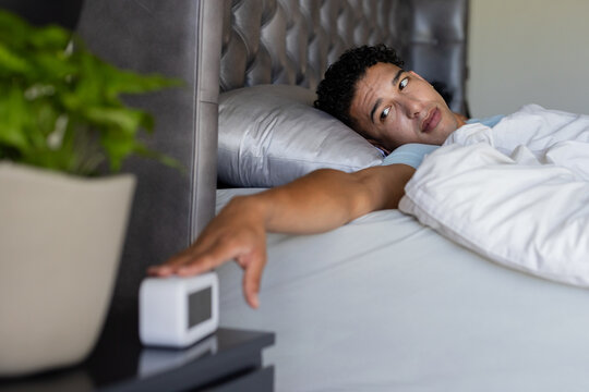 African American man lying in bedroom with light blue T shirt reaching for digital alarm clock