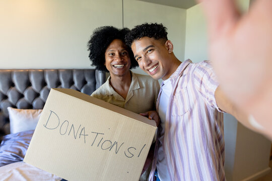 African American friends smiling, holding DONATIONS! box in casual shirts taking selfie in bedroom