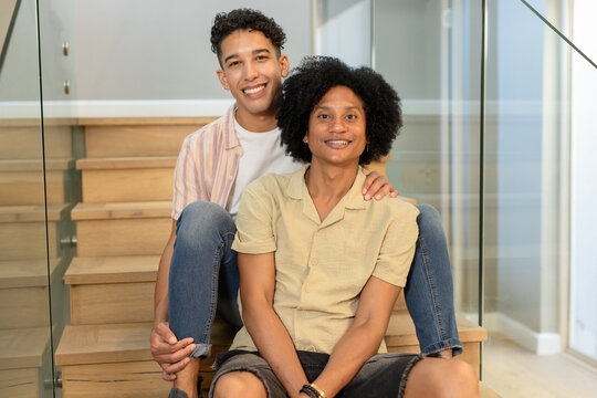 African American men sitting on wooden stairs at home, smiling with glass railing and bracelet