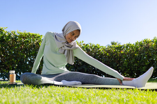 Woman wearing hijab, athletic wear stretching on mat in park, reaching to foot near reusable bottle