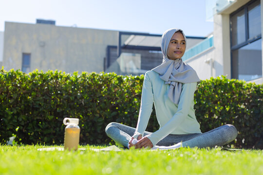 Adult woman wearing hijab and activewear sitting on yoga mat on lawn stretching with water bottle