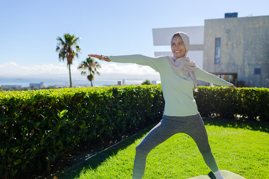 Woman standing in yoga pose on yoga mat on lawn, wearing sportswear headscarf, copy space