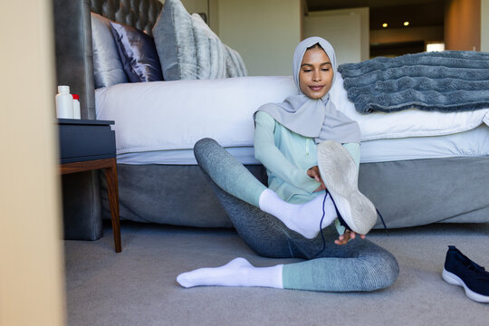 Adult woman in gray hijab and mint-green top tying shoe on bedroom floor by tufted headboard