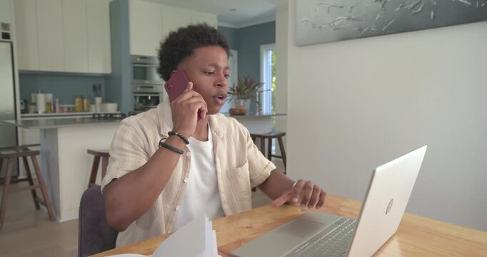 Adult male taking work call, holding magenta phone, typing on laptop at dining table with papers