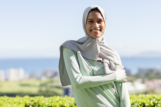 Female smiling, standing wearing light grey headscarf and pale green top by hedge overlooking sea