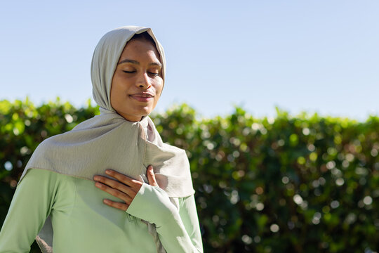 Adult African woman posing hand on chest eyes closed gray-hijab mint-top hedge blue-sky, copy space