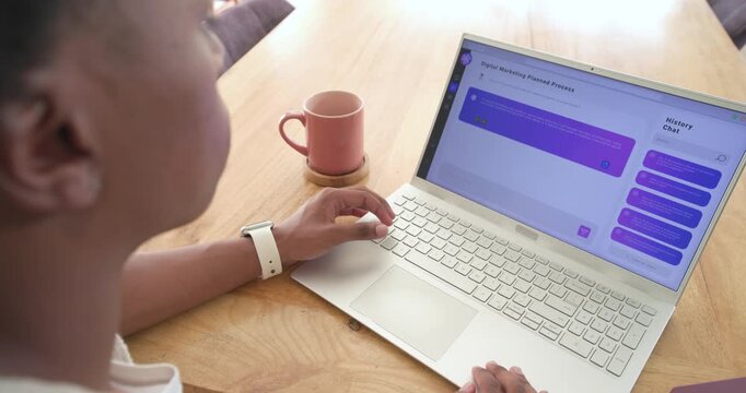 African man in 20s typing, reading chat appearing on laptop at wooden table, pointing, copy space