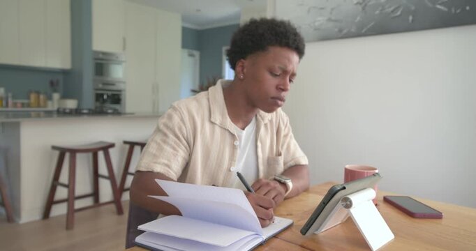 African American man 20s studying at wooden table consulting tablet and writing in stacked notebook