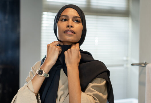 Woman adjusting black hijab, checking fit in bathroom by blinds, wearing beige top and watch