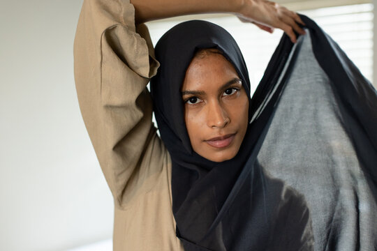 Woman wearing beige top and black headscarf draping fabric at home by window blinds