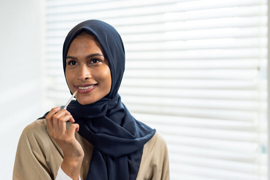 African woman applying lip gloss with wand, wearing navy hijab and beige outer garment by blinds