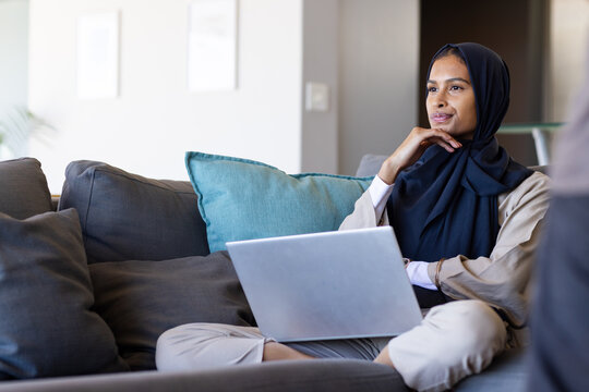 Woman in hijab sitting on sectional with teal cushion holding silver laptop at home, copy space