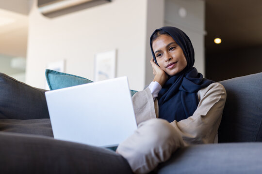 African American woman sitting on dark gray sofa using white laptop at home, wearing navy hijab
