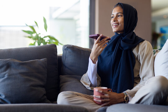 African-American woman in hijab sitting on sofa at home speaking into phone with mug, copy space