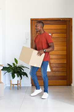 Adult man wearing red tee and jeans carrying cardboard box while standing in entryway beside plant