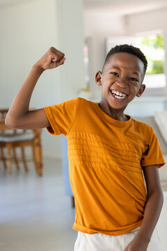 Male child smiling and flexing left arm in living area wearing orange t-shirt near wooden table