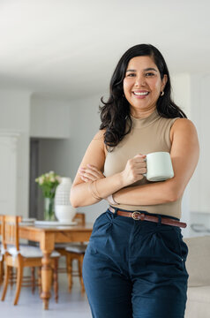 Mid adult woman standing in contemporary home holding pale ceramic mug, wearing navy trousers