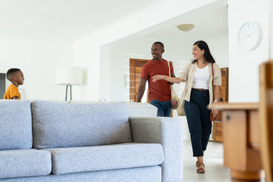 Diverse family entering bright living room with light-gray sofa, woven tote, shoulder bag