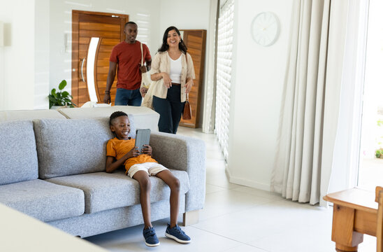 Diverse family entering home while child lounging on gray sectional sofa holding tablet, copy space