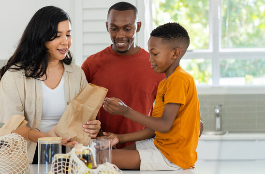 Diverse family unpacking groceries at kitchen island, woman holding brown paper bag, boy reaching