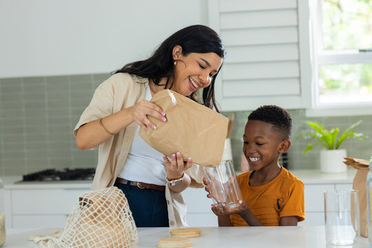 Mid adult woman pouring paper bag contents while youth boy holding clear jar on countertop smiling