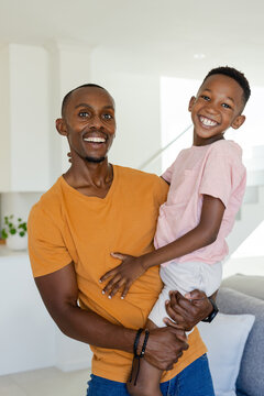 Adult father holding youth son at home near potted plant, smiling toward camera wearing wristwatch