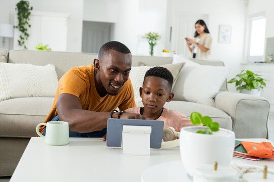 African American father and son leaning over tablet at living room coffee table, headphones nearby
