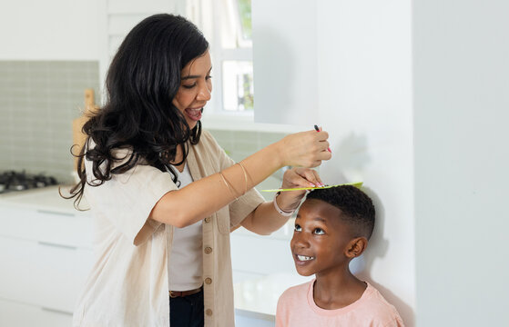 Adult woman beige shirt measuring child at white kitchen wall, marking height with yellow ruler