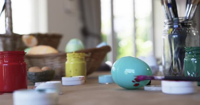 Paintbrush is leaving magenta smear on turquoise egg on wooden table near window, decorating eggs