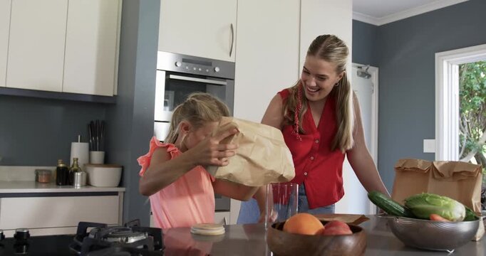 Woman guiding girl examining brown grocery bag, tipping contents into tall glass at kitchen island