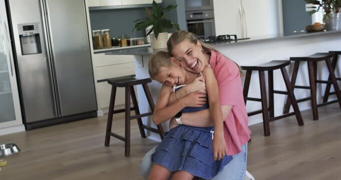 Mother wrapping arms around daughter in playful hug, laughing, squirming on stool at kitchen island
