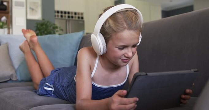 Preteen girl tapping play on tablet with headphones on sofa at home watching for fun laughing