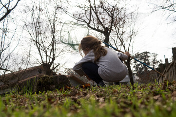 A wide shot from a low perspective showing a child crouching in the grass with a butterfly net against a bright sky and bare trees.