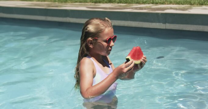 Child in swimsuit with heart shades holding watermelon slice, inspecting before biting in home pool