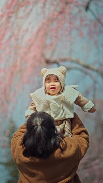A joyful Asian mother lifting her adorable toddler daughter high in the air under blooming pink cherry blossoms during a sunny spring day, capturing a heart-touching moment of pure happiness, family